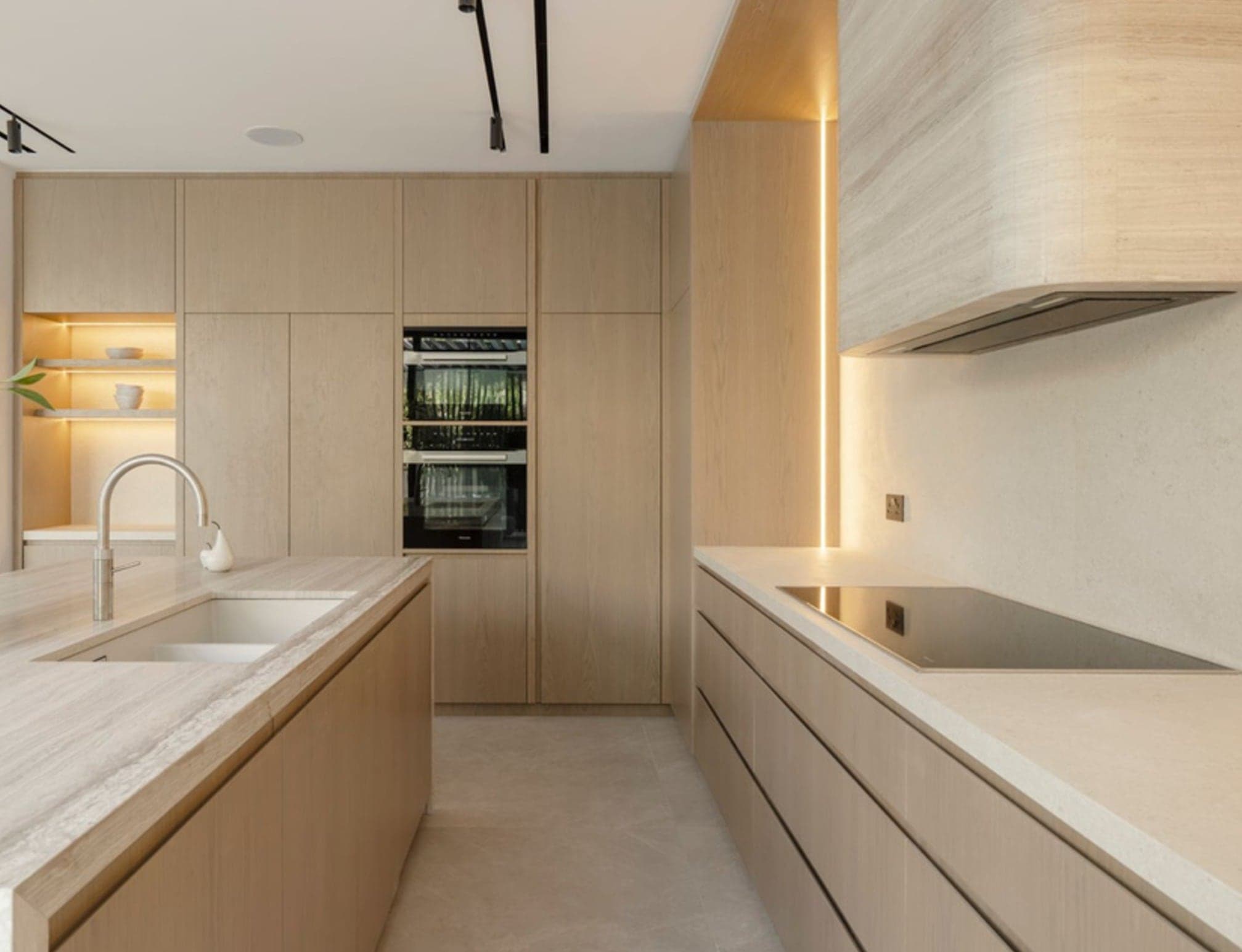Galley kitchen run in pale oak with a travertine worktop and integrated ovens